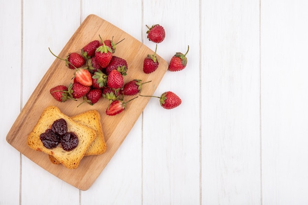 Top view of fresh strawberries on a wooden kitchen board with toasted bread on a white wooden background with copy space