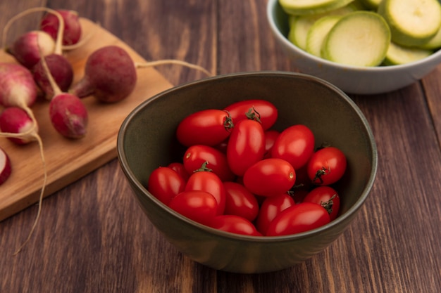 Top view of fresh tomatoes on a bowl with radishes on a wooden kitchen board with chopped zucchinis on a bowl on a wooden surface