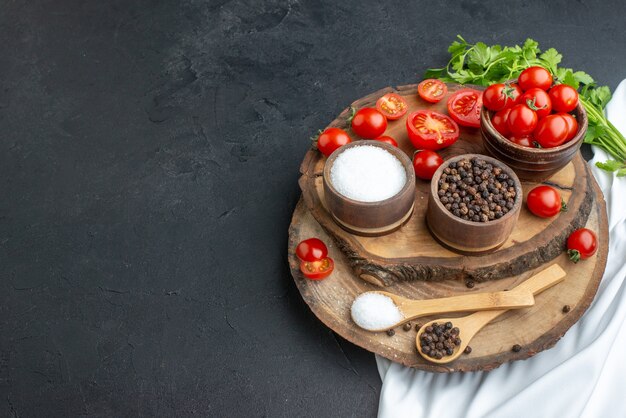 Top view of fresh tomatoes and spices in bowls spoons on wooden board on the left side on black surface