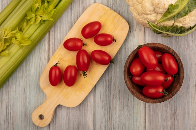 Top view of fresh tomatoes on a wooden kitchen board with tomatoes on a wooden bowl with cauliflower and celery isolated on a grey wooden surface