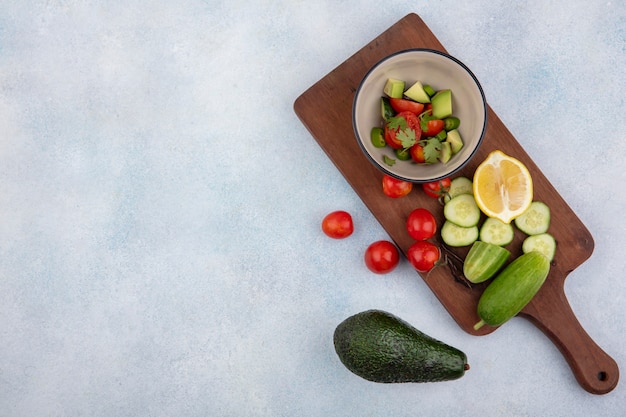 Top view of fresh vegetables including cucumber and tomato in a bowl on wood kitchen board with chopped cucumber slices cherry tomatoes lemonnd avocado on white