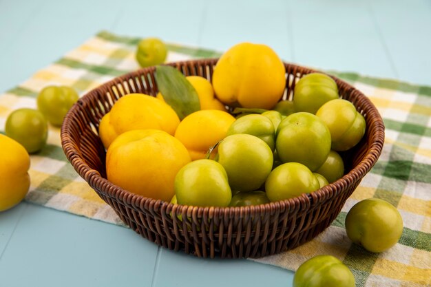 Top view of fresh yellow peaches on a bucket with green cherry plums on a checked cloth on a blue background