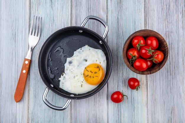 Free photo top view of fried egg in pan with fork and bowl of tomato on wood