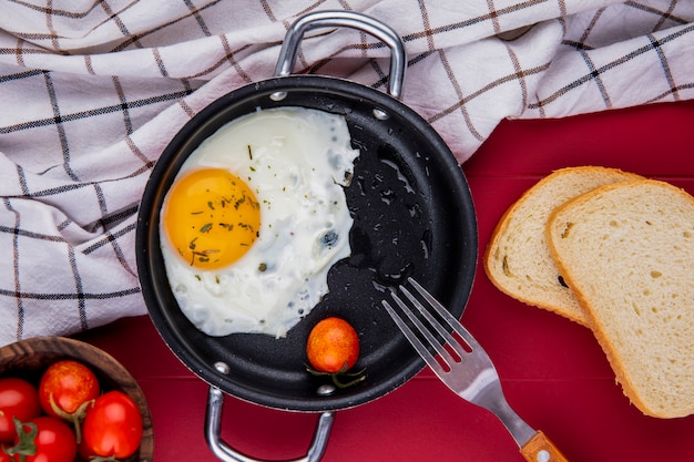 Free photo top view of fried egg with tomato and fork in pan on plaid cloth and bread slices bowl of tomatoes on red