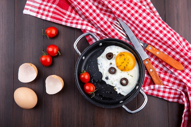 Free photo top view of fried egg with tomatoes and olives in pan and fork with knife on plaid cloth with egg and egg shell with tomatoes on wood