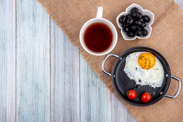 Top view of fried egg with tomatoes in pan and cup of tea with bowl of black olive on sackcloth and wood with copy space