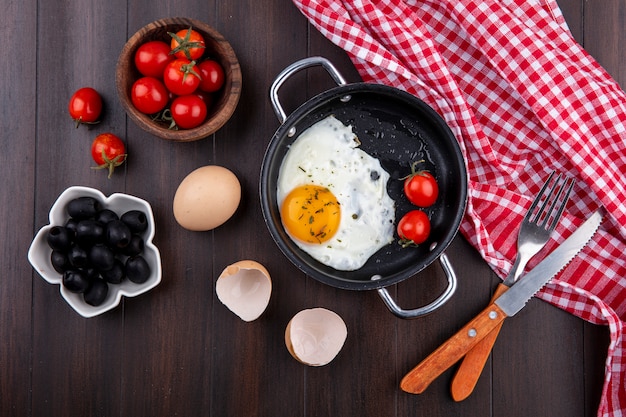 Free photo top view of fried egg with tomatoes in pan and fork with knife on plaid cloth and egg with shell and bowls of tomato and olive on wood