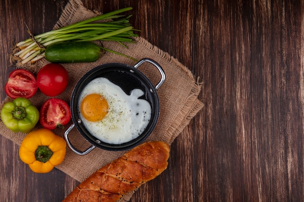 Free photo top view fried eggs in a pan with green onions  tomatoes  cucumber  bell peppers and a loaf of bread  on a beige napkin  on a wooden background