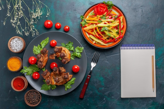 Top view fried meat slices with sliced vegetable salad notepad and seasonings on dark-blue desk vegetable food meat health meal