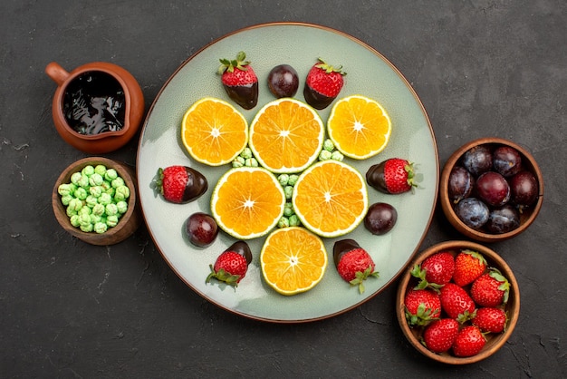 Top view from afar berries and chocolate plate of chopped orange and chocolate-covered strawberries next to the bowls with sweets berries nd chocolate sauce