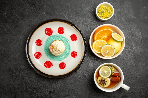 Top view from afar berries and chocolate plate with cupcake with white cream and sauces next to the bowls of herbs and sliced lemons and cup of tea