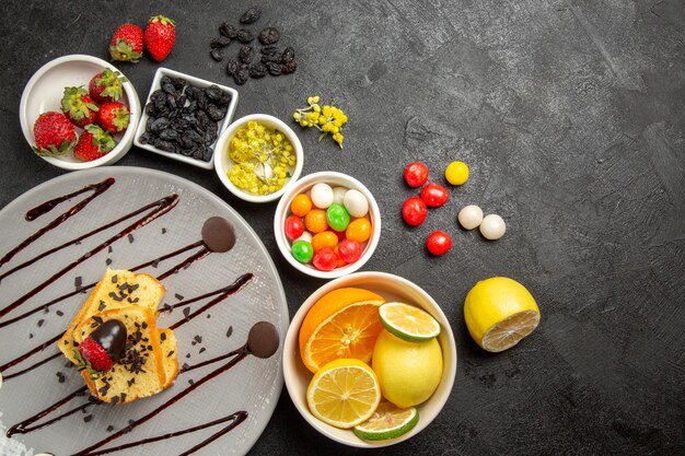 Top view from afar berries and fruits cake with chocolate-covered strawberries next to the white bowls of strawberries limes lemons oranges and colorful sweets on the table