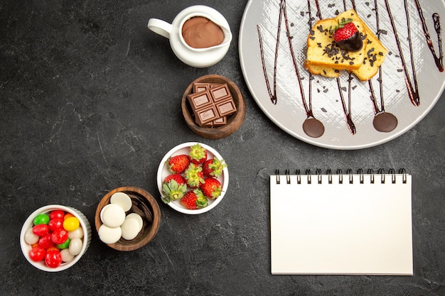 Free Photo top view from afar cake with strawberries plate of cake with strawberries next to the bowls of sweets chocolate strawberries and chocolate cream and white notebook