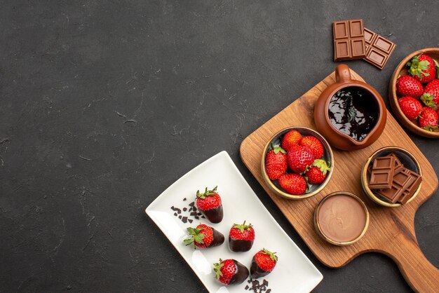 Top view from afar chocolate on board chocolate-covered strawberries on plate next to the cutting board with chocolate cream and strawberries and bars of chocolate