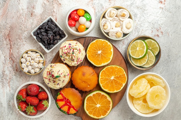 Top view from afar citrus fruit on the board bowls of berries sweets and dried pineapples and sliced orange and cookies on the wooden cutting board