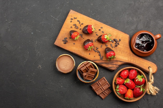 Free Photo top view from afar cutting board chocolate-covered strawberries on cutting board next to chocolate cream and strawberries on the right side of the dark table