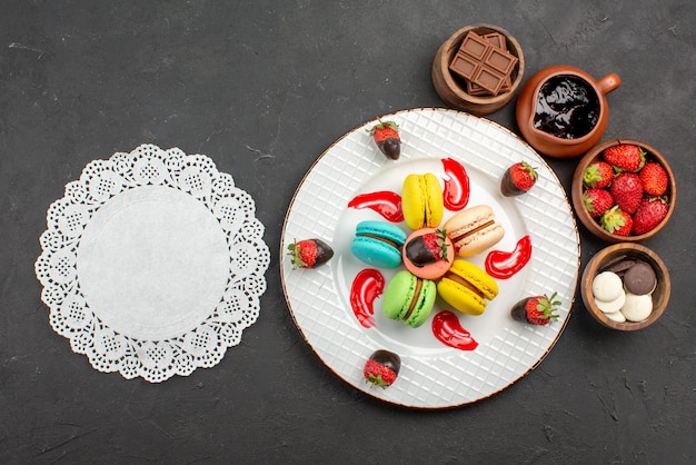 Top view from afar dessert macaroons and strawberries in the plate next to the lace doily and bowls with chocolate strawberries and chocolate cream on the table