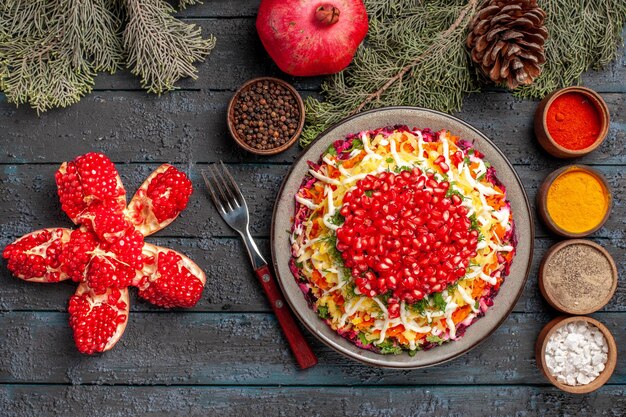 Top view from afar dish and tree branches appetizing dish of pomegranate next to the bowls of spices fork peeled pomegranate and tree branches with cones on the dark table