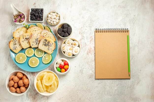 Top view from afar lemon cake seven bowls of candies next to the cream notebook with green pencil plate of pieces of cake with lemon on the table