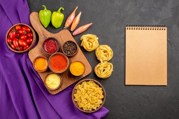 Free Photo top view from afar pasta and tomatoes spices and sauces in bowls on the cutting board onion bowl of tomatoes ball pepper and pasta next to the cream notebook on the table