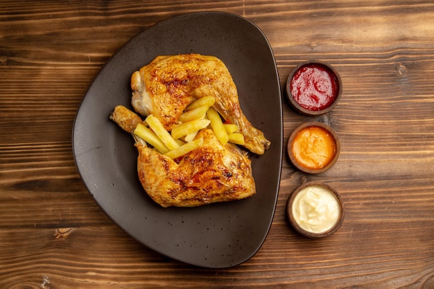 Top view from afar plate of fast food bowls of colorful sauces next to the plate of appetizing french fries and chicken legs on the wooden table