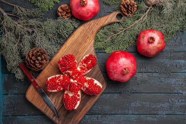 Top view from afar pomegranate on board red pilled pomegranate on cutting board next to ripe three pomegranates knife and spruce branches with cones on table