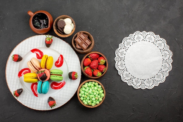 Top view from afar strawberries and macaroons plate of appetizing French macaroons and strawberries bowls of sweets around it and lace doily on the table