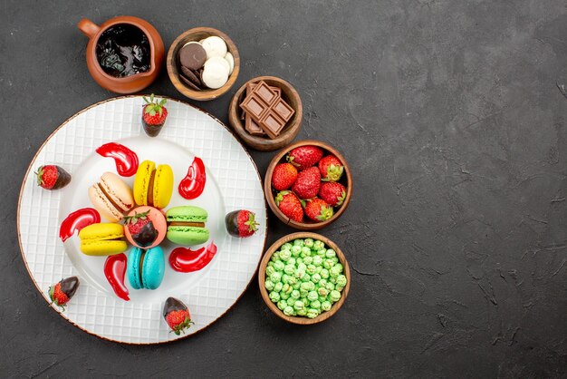 Top view from afar strawberries and macaroons plate of appetizing French macaroons and strawberries and bowls of sweets around it on the table