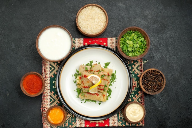 Free photo top view from afar stuffed cabbage stuffed cabbage and bowls of herbs white and yellow sauces black pepper spices rice and sour cream on multicolored checkered tablecloth on dark surface