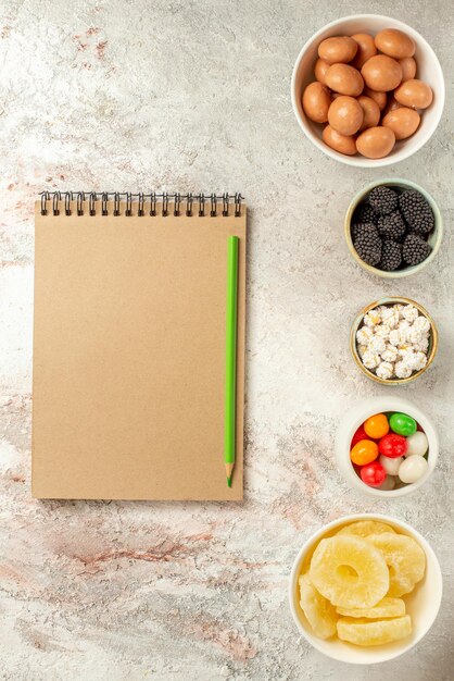 Top view from afar sweets in bowls appetizing sweets in bowls next to the green pencil and cream notebook on the light surface