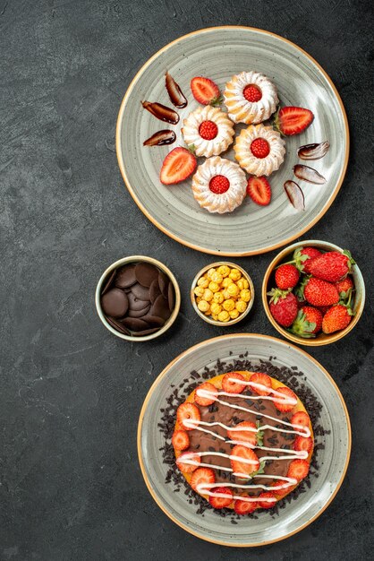 Top view from afar sweets and cake strawberry cookies and cake with chocolate and bowls of strawberry hizelnuts chocolate on the right side of black table