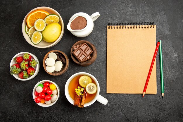 Top view from afar sweets on the table a cup of tea with cinnamon and lemon next to the cream notebook with green and red pencil and bowls of strawberries chocolate and candies