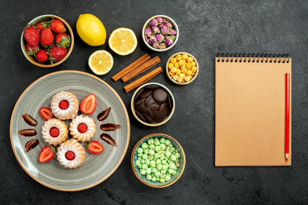 Free photo top view from afar sweets with tea cookies with strawberry black tea with lemon hizelnuts bowls of chocolate and different sweets next to cream notebook and red pencil on dark surface