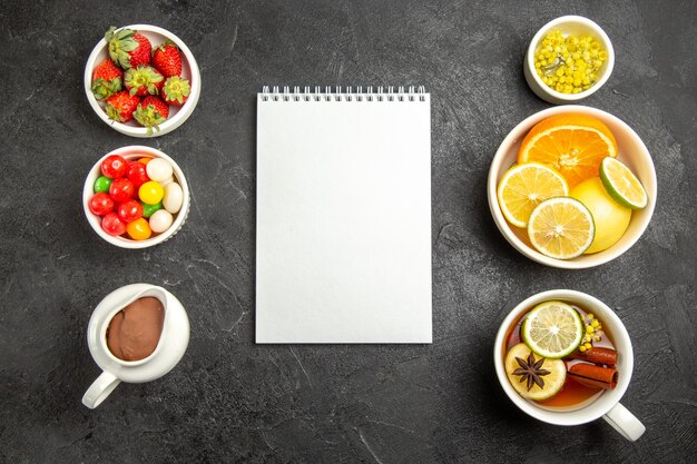 Top view from afar sweets with tea a cup of tea with cinnamon and star anise next to the white notebook and the bowls of herbs citrus fruits chocolate cream strawberries on the table