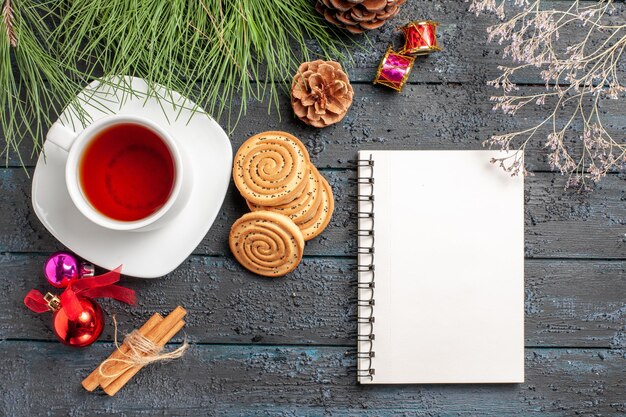 Top view from afar tea spruce tree with cones and Christmas toys next to the cinnamon sticks a cup of tea on the saucer and white notebook