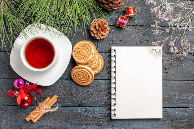 Top view from afar tea spruce tree with cones and Christmas toys next to the cinnamon sticks a cup of tea on the saucer and white notebook