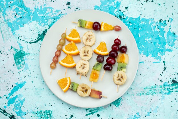 Top view fruit composition sliced on sticks inside white plate on the bright desk fruit exotic cookie sugar