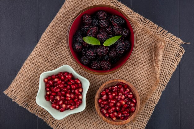 Top view of fruits as blackberry and pomegranate berries in bowls on sackcloth and black surface