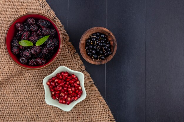 Top view of fruits as blackberry and pomegranate berries in bowls on sackcloth with bowl of sloe berries on black surface