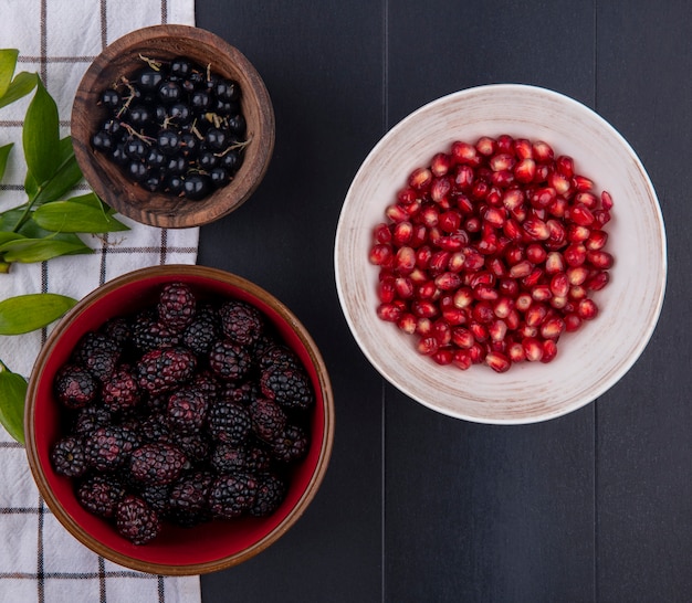 Top view of fruits as bowls of sloe and blackberry with leaves on plaid cloth and bowl of pomegranate berries on black surface