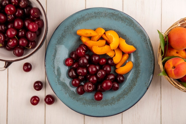 Top view of fruits as cherries and apricot slices in plate and basket of apricots with bowl of cherries on wooden background