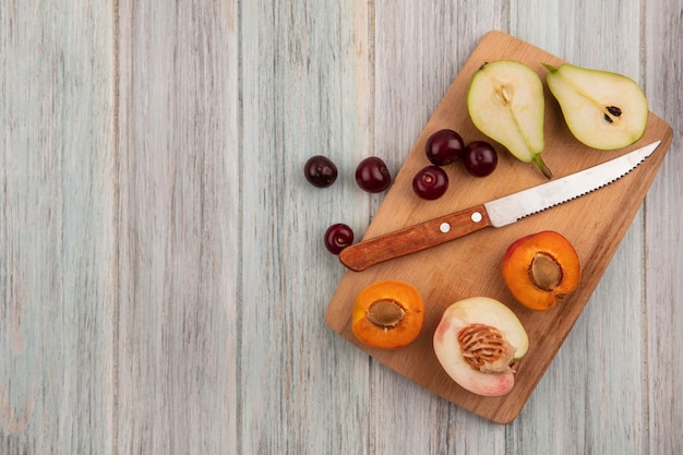 Top view of fruits as cherries and half cut apricot pear and peach with knife on cutting board on wooden background with copy space