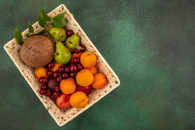 Top view of fruits as coconut peach apricot pear cherry with leaves in basket on green background with copy space