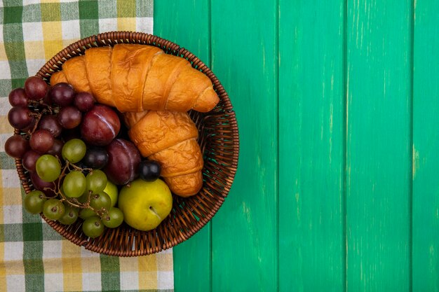 Top view of fruits as grape pluots sloe berries with croissants in basket on plaid cloth and green background with copy space