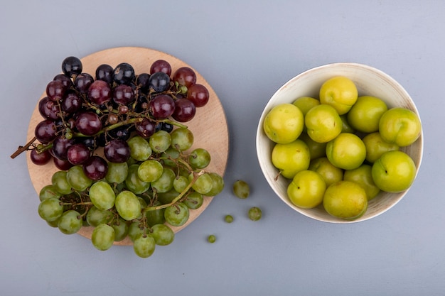 Free photo top view of fruits as grapes on cutting board and bowl of plums on gray background