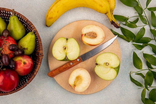 Top view of fruits as half cut peach and apple with knife on cutting board and basket of pear grape peach with banana and leaves on white background