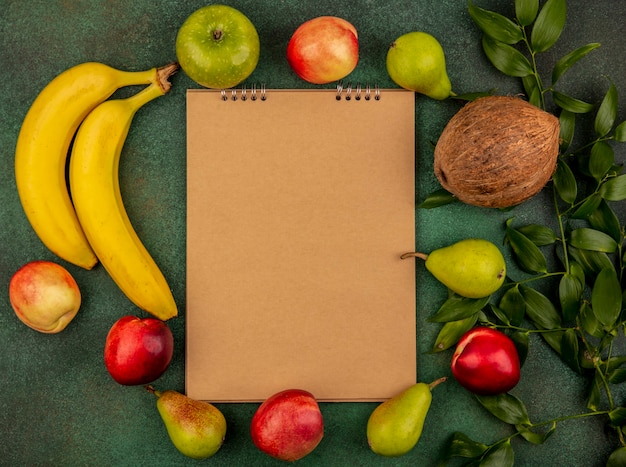 Free photo top view of fruits as peach coconut apple pear banana with leaves around note pad on green background with copy space