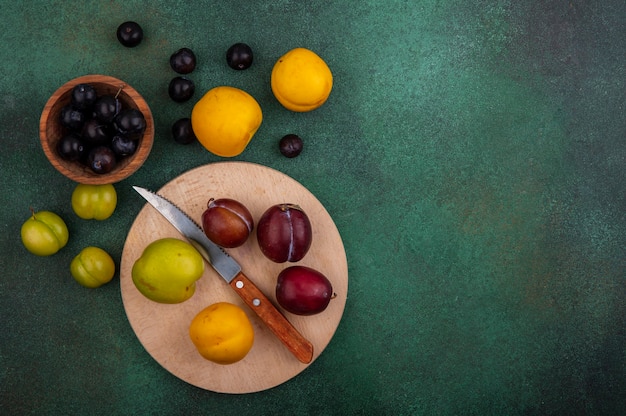 Free photo top view of fruits as pluots and nectacot with knife on cutting board and grape berries in bowl with plums nectacots and grape berries on green background with copy space