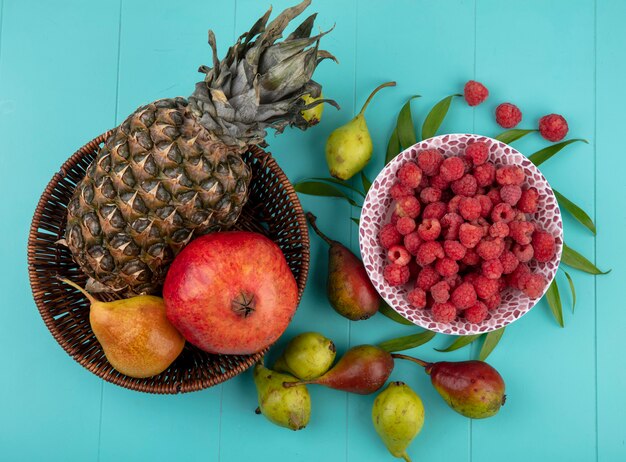 Top view of fruits in basket and bowl of raspberry with peaches and leaves on blue surface