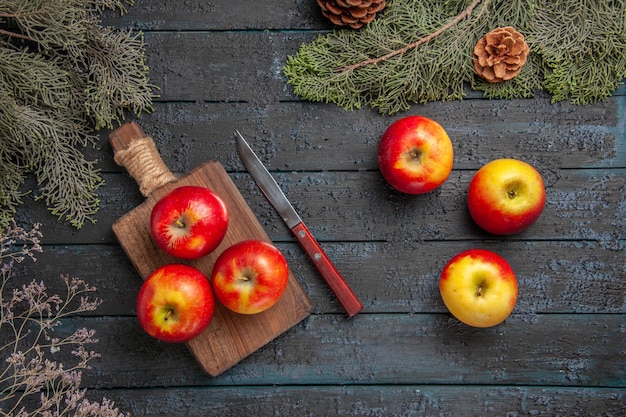 Free Photo top view fruits and knife three yellow-reddish apples on the wooden cutting board next to a knife and three apples under the tree banches with cones on the table